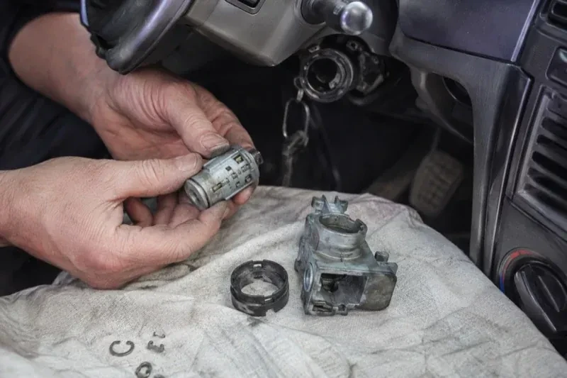 Hands holding a removed ignition cylinder beside a car steering column, with the housing and small parts on a cloth. — Ignition Lock Repair, London